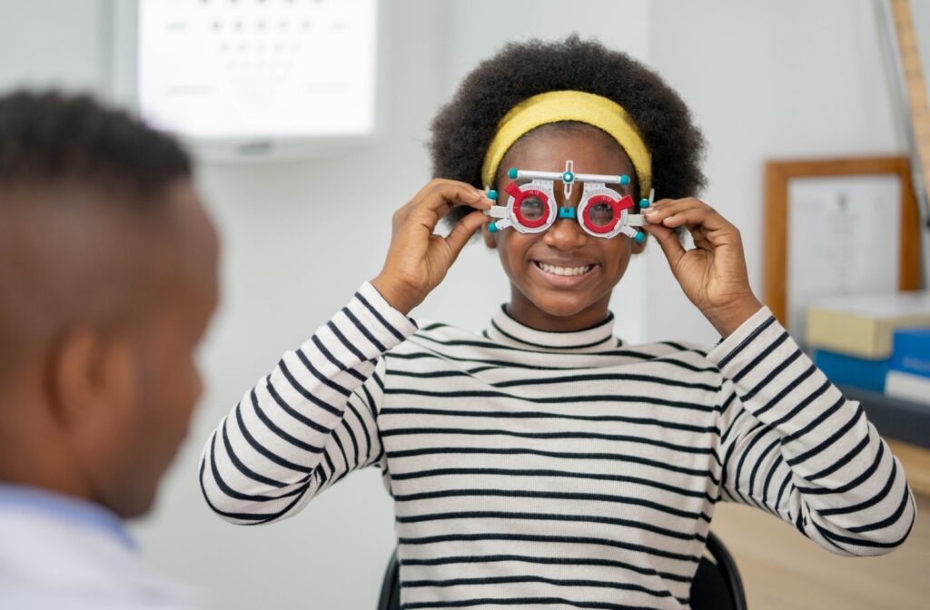 A young eye patient holds a colourful glasses-like device to her eyes and smiles at her optometrist.