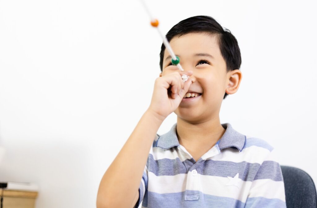 A young boy in a striped shirt smiles as he holds a Brock string up to his eye as he performs a vision therapy exercise