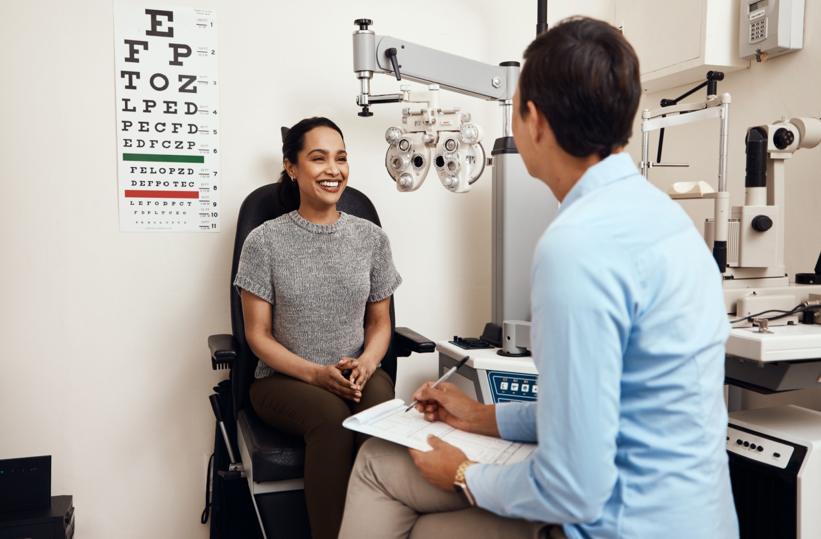 A young woman smiles at her optometrist while in the office for an eye test for vision therapy, an eye chart on the wall