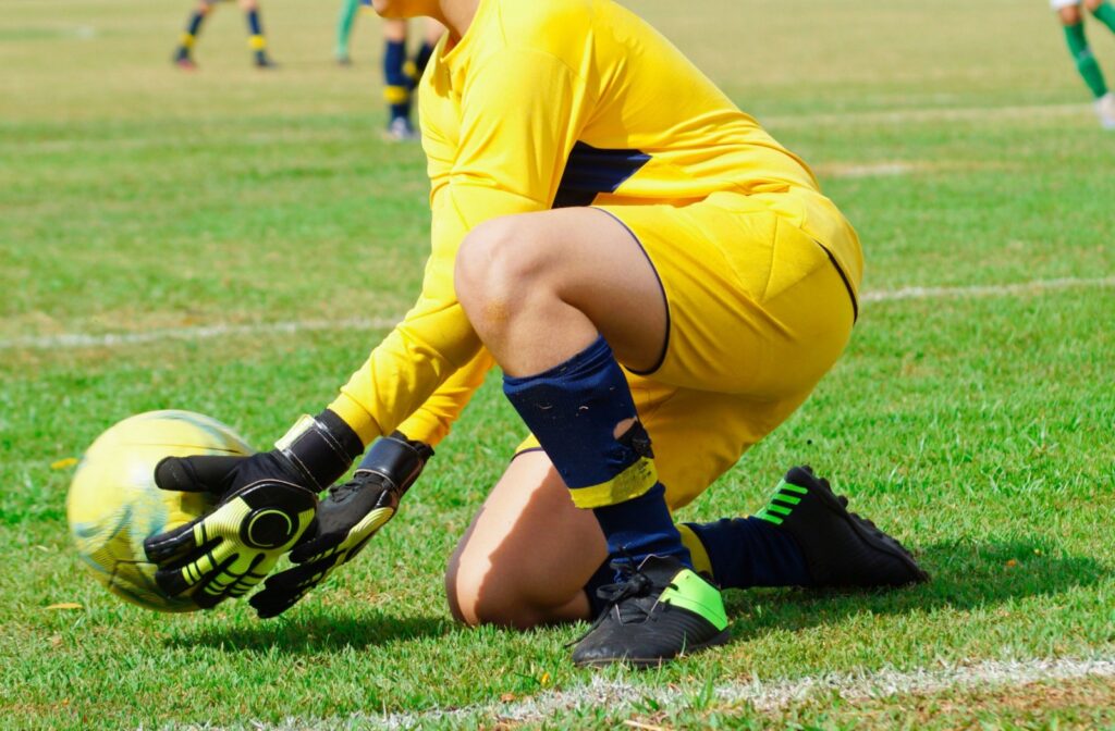 A goalkeeper in a yellow jersey bends down to stop a soccer ball headed towards their net.