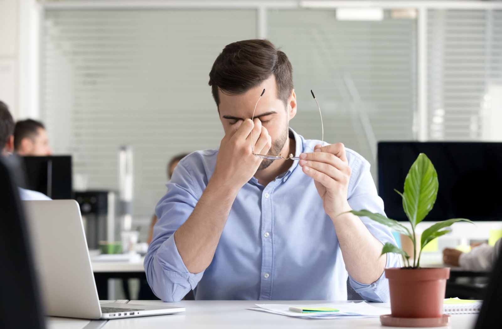 A young adult taking off their glasses at work to rub their eyes in discomfort, wondering if they need vision therapy.