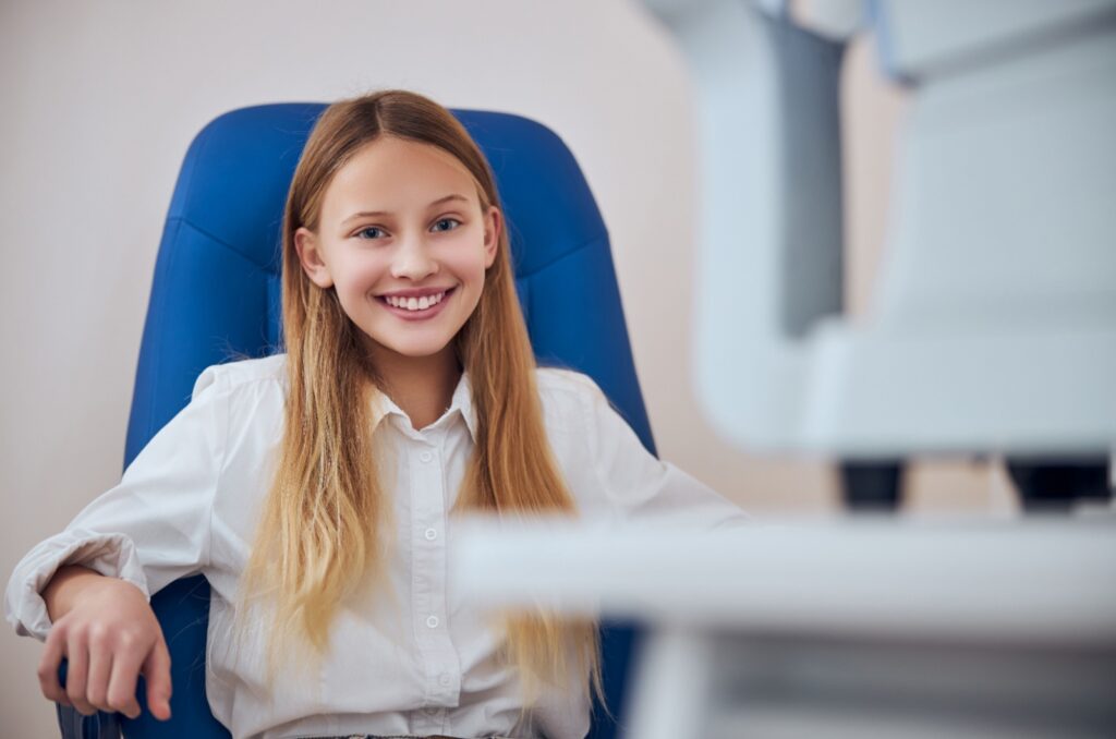 A smiling child sitting in a blue chair waiting for an eye exam.