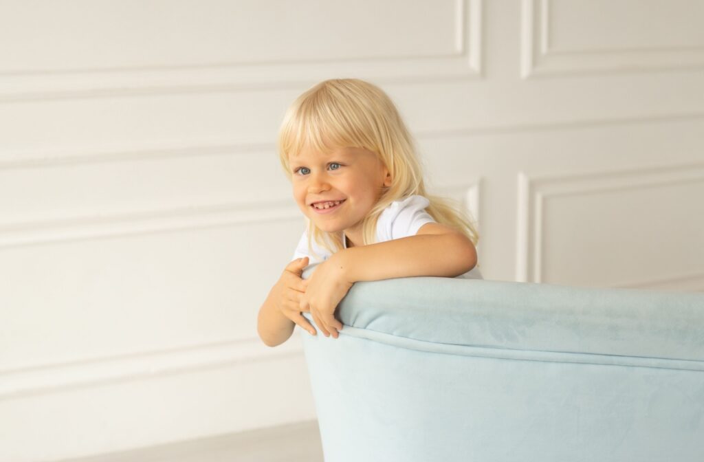 Young child with long blond hair and crossed eyes leaning over the back of a light turquoise couch.